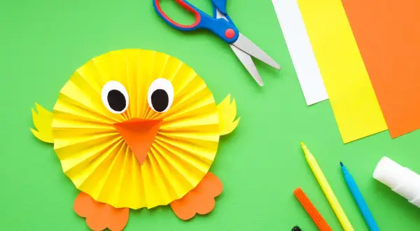 Un pollito de papel, ideal para manualidades fáciles para niños de 3 a 6 años. Tijeras, rotuladores y cartulinas de colores completan la escena.