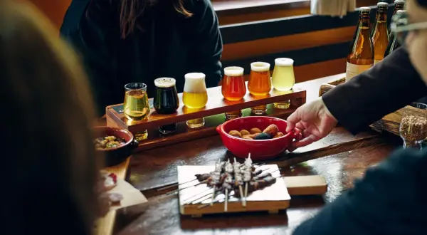 Degustazione di birre pregiate: un assortimento di diverse birre in bicchierini, accompagnate da stuzzichini e spiedini su un tavolo di legno.