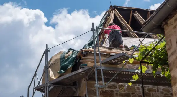 Un ouvrier travaille sur un toit en rénovation, une étape essentielle pour garantir ma prime logement décent. Le ciel bleu et les nuages blancs surplombent le chantier.