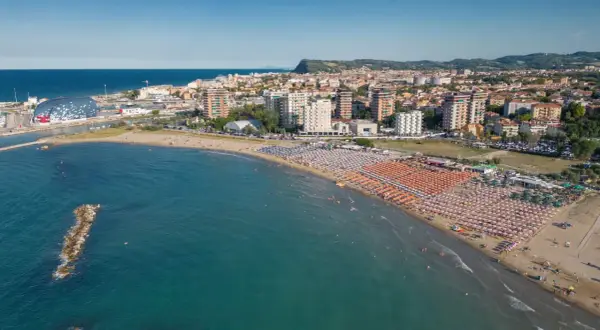 Vista aerea della Pesaro strand, con ombrelloni colorati, mare azzurro e la città sullo sfondo.
