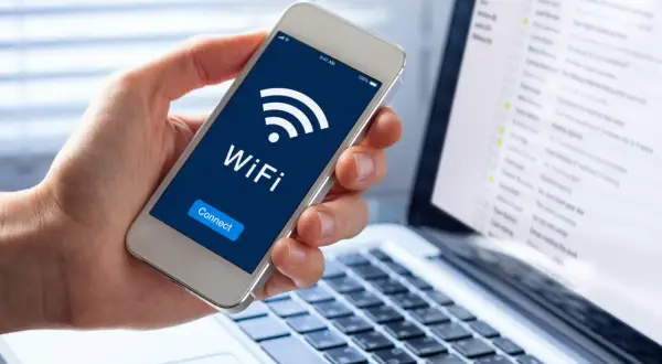 Dłoń trzyma smartfon z ikoną WiFi i napisem "Connect". Obok leży laptop z widokiem skrzynki mailowej. Dostęp do sieci, być może przez **ssid**, jest kluczowy.