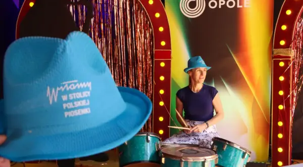 Kobieta w niebieskim kapeluszu gra na perkusji przed sceną z napisem "Opole".