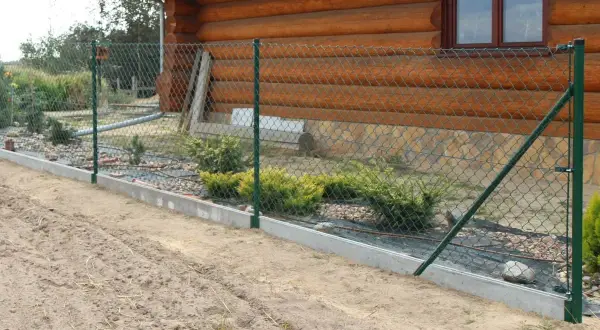 Zielona siatka ogrodzeniowa, zamontowana na betonowym podmurówce, otacza drewniany dom. Wokół rosną krzewy.