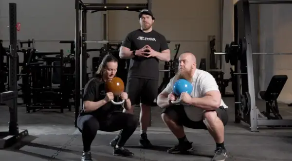 Två personer utför goblet squat med kettlebells medan en instruktör står bakom.