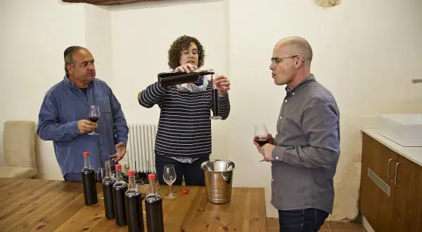 Weinprobe: Eine Frau gießt Wein ein, während zwei Männer zuschauen. Flaschen und Gläser stehen bereit für die assemblage wein.