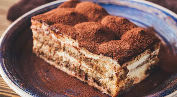 Une part de tiramisu, saupoudrée de cacao, sur une assiette bleue. Un délice qui évoque tous les ingrédients du tiramisu.