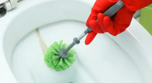 Mano con guanto rosso pulisce il calcare dal wc con uno scopino verde.