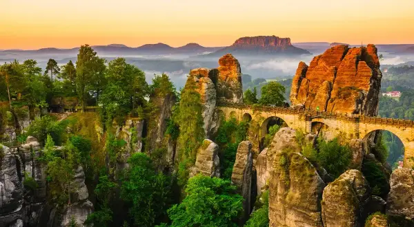 Un pont de pierre traverse des formations rocheuses spectaculaires, rappelant la Suisse bohémienne. La nature y est luxuriante, baignée par la lumière dorée du crépuscule.