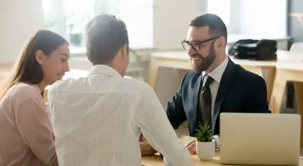 Coppia sorridente discute con un consulente di surroga mutuo consap, con laptop e pianta sul tavolo.