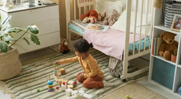 Ein kleines Kind spielt mit Holzbausteinen in einem gemütlichen, quadratischen Kinderzimmer. Spielzeug und ein Bett laden zum Verweilen ein.