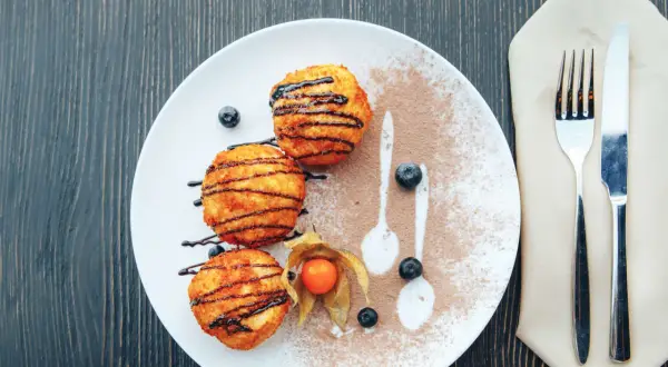 Drei goldbraune, knusprige Kugeln, beträufelt mit Schokoladensauce, liegen auf einem Teller mit Kakaopulver und Beeren. Ein süßer Genuss, der an Eis erinnert, aber als knusprige Leckerei serviert wird.