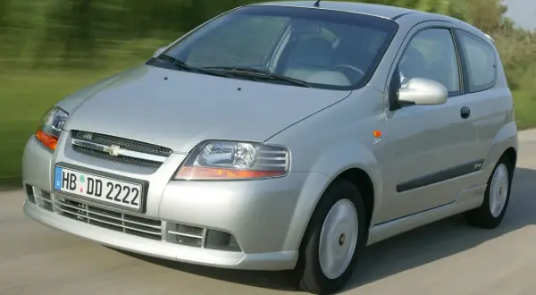 Srebrny Chevrolet Kalos w wersji 3-drzwiowej jedzie po drodze, otoczony zielenią.