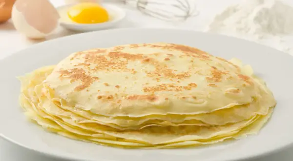 Ein Stapel goldbrauner Palatschinken auf einem Teller, daneben Eier, ein Schneebesen und Mehl. Perfekt für ein Palatschinken Rezept!