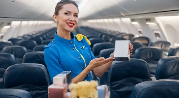 Uśmiechnięta stewardessa w niebieskim mundurku prezentuje pudełko z perfumami do samolotu.