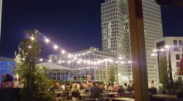 Abendstimmung auf einer Dachterrasse in Berlin. Lichterketten beleuchten Tische, an denen Gäste sitzen. Im Hintergrund ragen moderne Gebäude auf, die an den Spreegold Bikini erinnern.