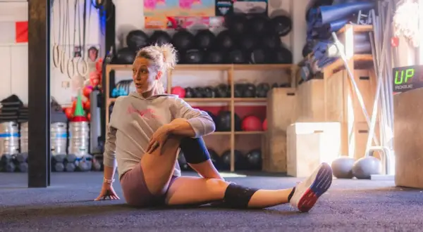 Femme en tenue de sport effectuant un exercice de mobilité au sol, dans une salle de sport avec équipement en arrière-plan.