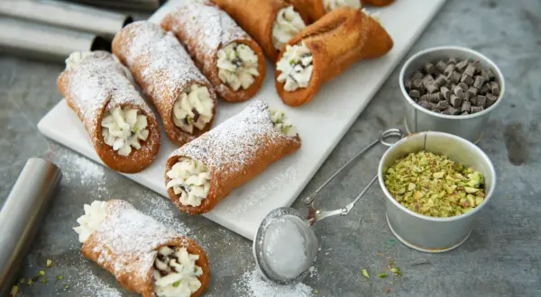 Färska cannoli med krämig fyllning, pudrade med florsocker. Ingredienser för ett perfekt cannoli recept syns bredvid.