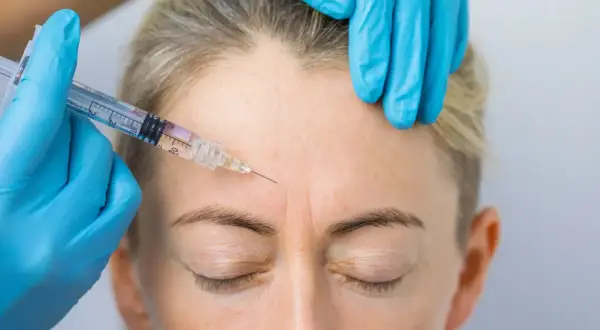 Zabieg baby botox na czole. Ręka w niebieskiej rękawiczce trzyma strzykawkę z igłą, która wstrzykuje preparat w zmarszczki mimiczne.