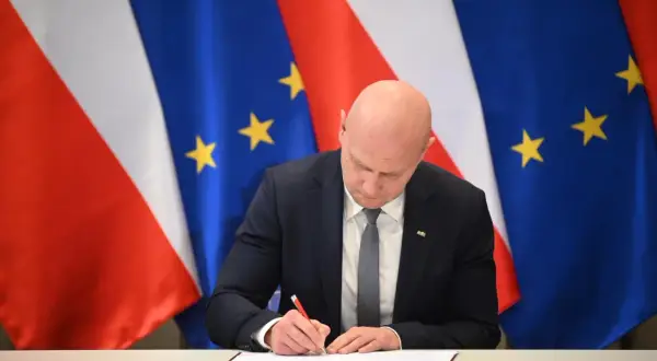 Łysy mężczyzna w garniturze podpisuje dokument na tle flag Polski i UE. Spodziewaj się niespodziewanego, bo nigdy nie wiadomo, co przyniesie przyszłość.