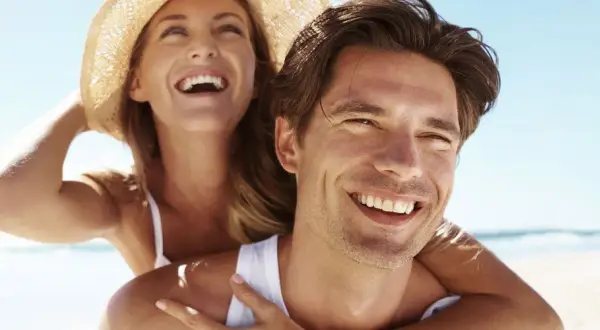 Glückliches Paar am Strand, die Frau lacht und trägt einen Hut. Selbst im Schatten wird man braun, wenn man die Sonne genießt.