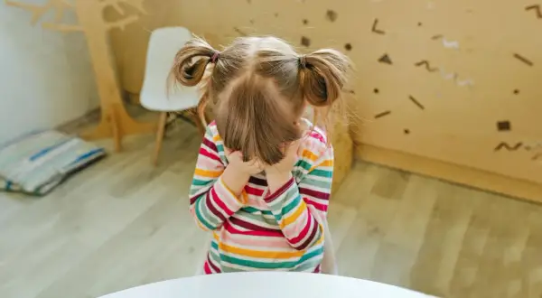 Mała dziewczynka w kolorowym sweterku, z włosami spiętymi w dwa kucyki, zakrywa twarz dłońmi. Wygląda jak karny jeżyk, który chce się schować przed całym światem.