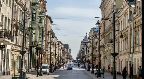 Belebte Straße in **Łódź, Polen**, gesäumt von historischen Gebäuden und Straßenlaternen. Autos und Fußgänger beleben die Szene.