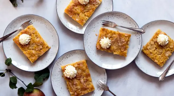 Saftiger Apfelkuchen vom Blech, garniert mit Sahnetuffs und Mandelblättchen, auf Tellern angerichtet.