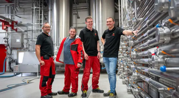Vier Männer in Arbeitskleidung stehen vor großen Edelstahltanks und Rohrleitungen. Sie sind Teil einer Ausbildung zum Brauer und blicken stolz in die Kamera.