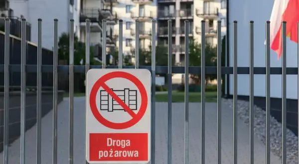 Znak zakazu wjazdu na drogę pożarową na metalowej bramie. W tle budynki mieszkalne i polska flaga.