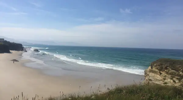 Un paseo solitario por las **playas de Lugo**, con el mar turquesa rompiendo suavemente en la arena dorada y acantilados rocosos.