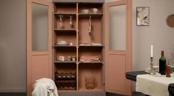 Une vieille armoire repeinte, aux portes roses et intérieur gris, abrite vaisselle, verres à pied, bouteilles de vin et paniers.