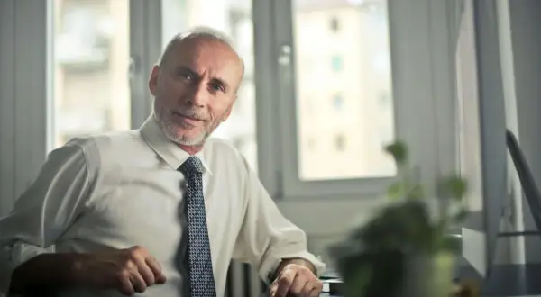 Starszy mężczyzna w białej koszuli i krawacie, z siwą brodą, siedzi przy biurku. Jego kod zawodu to pracownik administracyjny.