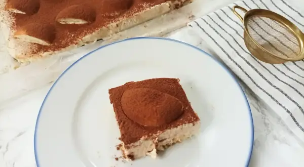 Pyszne ciasto cappuccino z mascarpone, posypane kakao, czeka na talerzu. Obok całe ciasto i sitko do posypywania.