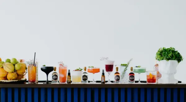 Rząd kolorowych drinków i butelek z alkoholami na barze, obok koszyka z owocami i białej rzeźby głowy.