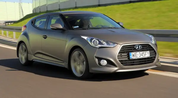 Szary Hyundai Veloster pędzi po drodze, za nim zielone ekrany dźwiękochłonne.