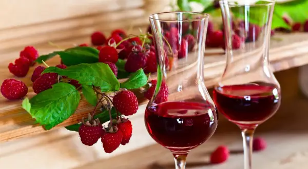 Dwa kieliszki z czerwonym winem malinowym, obok świeże maliny z liśćmi. Idealne na lato, jak zrobić wino z malin.