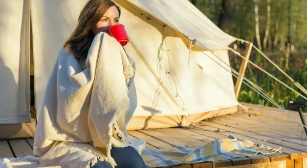 Kvinna med filt och röd mugg vid tältet. En mysig morgon som visar vikten av en bra packlista camping.