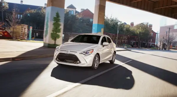 Biały Toyota Yaris Sedan jedzie pod wiaduktem, mijając budynki i drzewa.