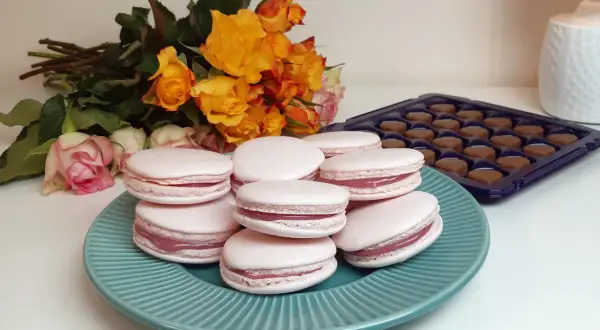 Różowe makaroniki z różowym nadzieniem, obok bukiet pomarańczowych i różowych róż oraz pudełko czekoladek. Idealne na prezent, z pysznym ganache do makaroników.