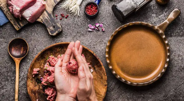 Mani che formano polpette di carne su un piatto di legno, con ingredienti freschi e una padella pronta.