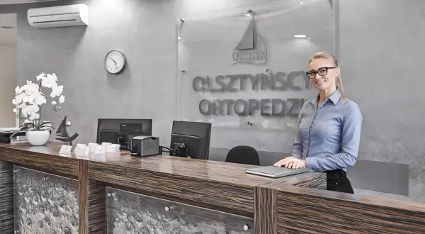 Recepcjonistka w okularach stoi przy ladzie w nowoczesnej klinice ortopedycznej.
