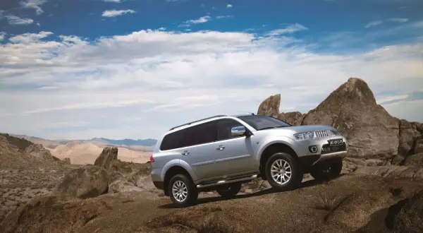 Srebrny Mitsubishi Pajero Sport pokonuje skalisty teren na tle pustynnego krajobrazu i błękitnego nieba z chmurami.