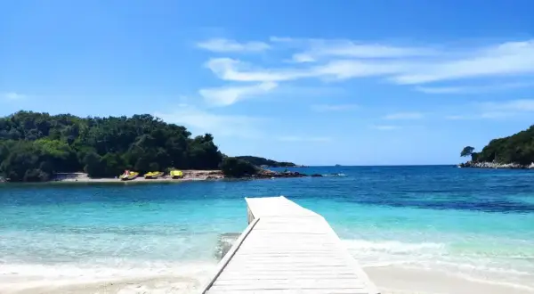 Drewniany pomost prowadzi do krystalicznie czystej wody na **Ksamil beach**. W oddali widać zielone wyspy i żółte łódki.