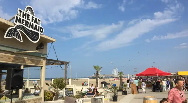 Strandpromenade in Den Haag mit dem Restaurant "The Fat Mermaid", Menschen, Sonnenschirmen und einem Riesenrad. Tolle Den Haag Unternehmungen!