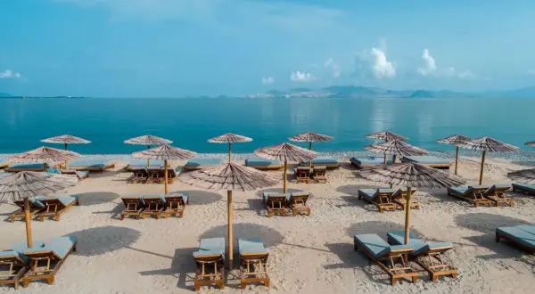 Puste leżaki i parasole na piaszczystej plaży, gotowe na gości. W tle spokojne, błękitne morze i górzyste wybrzeże. Idealne miejsce na relaks.