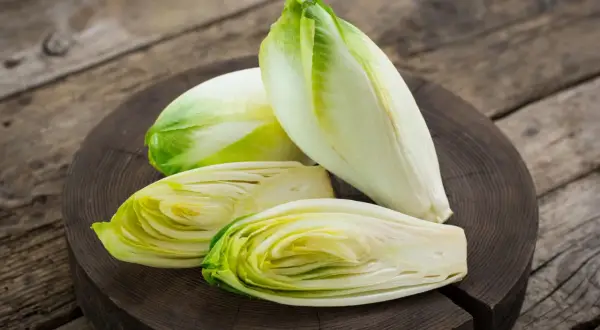 Frische Chicorée-Köpfe, einer davon halbiert, liegen auf einem Holzbrett. Perfekt für einen knackigen Chicorée-Salat.