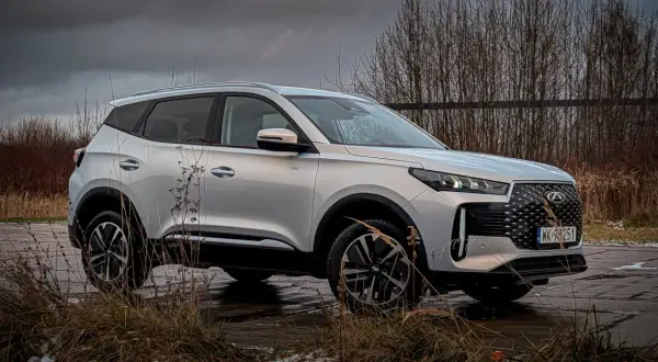 Srebrny Chery Tiggo 4 na tle ponurego krajobrazu. Auto stoi na mokrej nawierzchni, otoczone suchymi trawami i drzewami.