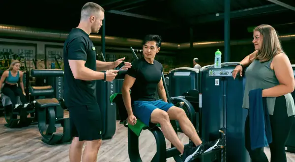 Trainer erklärt Mann die richtige Ausführung am Gerät für den erector spinae. Frau schaut zu.