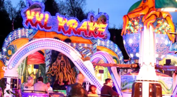 Bunte Lichter und Piratenmotive auf einer Kirmes in Berlin. Eine Achterbahn mit dem Schriftzug "Sky Krake" zieht Besucher an.
