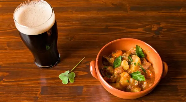 Un bicchiere di birra stout scura con schiuma cremosa accanto a una ciotola di stufato irlandese e un quadrifoglio su un tavolo di legno.